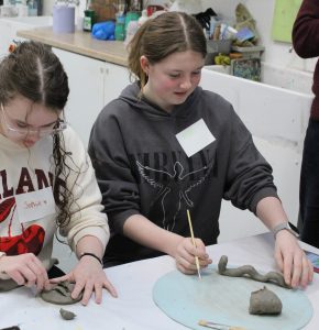 Two young girls are working on clay sculptures at a table in an art classroom. The girl on the left is wearing glasses and a cream-colored sweater with "ISLAND" written on it, and the girl on the right is wearing a gray hoodie with "Molly" on a name tag. They are focused on shaping their clay creations, with various art supplies and materials visible in the background.