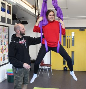 A young woman is sitting on purple aerial silk swings with her legs apart, smiling. She is wearing a red sweatshirt, black leggings, and white socks. A man with a beard, dressed in a black hoodie and camouflage shorts, is assisting or guiding her by holding the silk. They are in an indoor space with yellow double doors, a chair, and various equipment in the background.