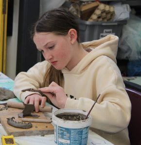 A young woman with long brown hair, wearing a beige hoodie, is focused on sculpting clay with a tool in a workshop or art classroom. She is sitting at a table with various clay pieces and sculpting tools, concentrating on her work. In the background, there are storage bins and supplies.