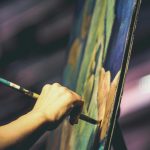 A close-up of a hand holding a stylus and drawing on a large digital display, which shows colorful abstract strokes against a dark background.
